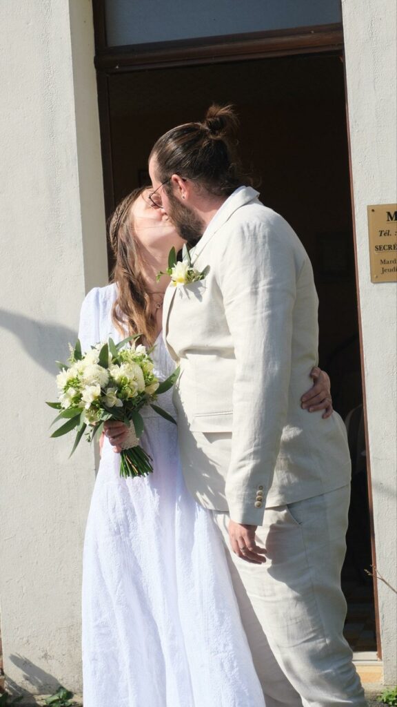 Un couple de mariés s’embrassant devant un bâtiment, la mariée tenant un bouquet de fleurs fraîches blanches et feuillage, et le marié portant une boutonnière assortie créée sur mesure par Invitation Florale créatrice située en Normandie près de Caen
