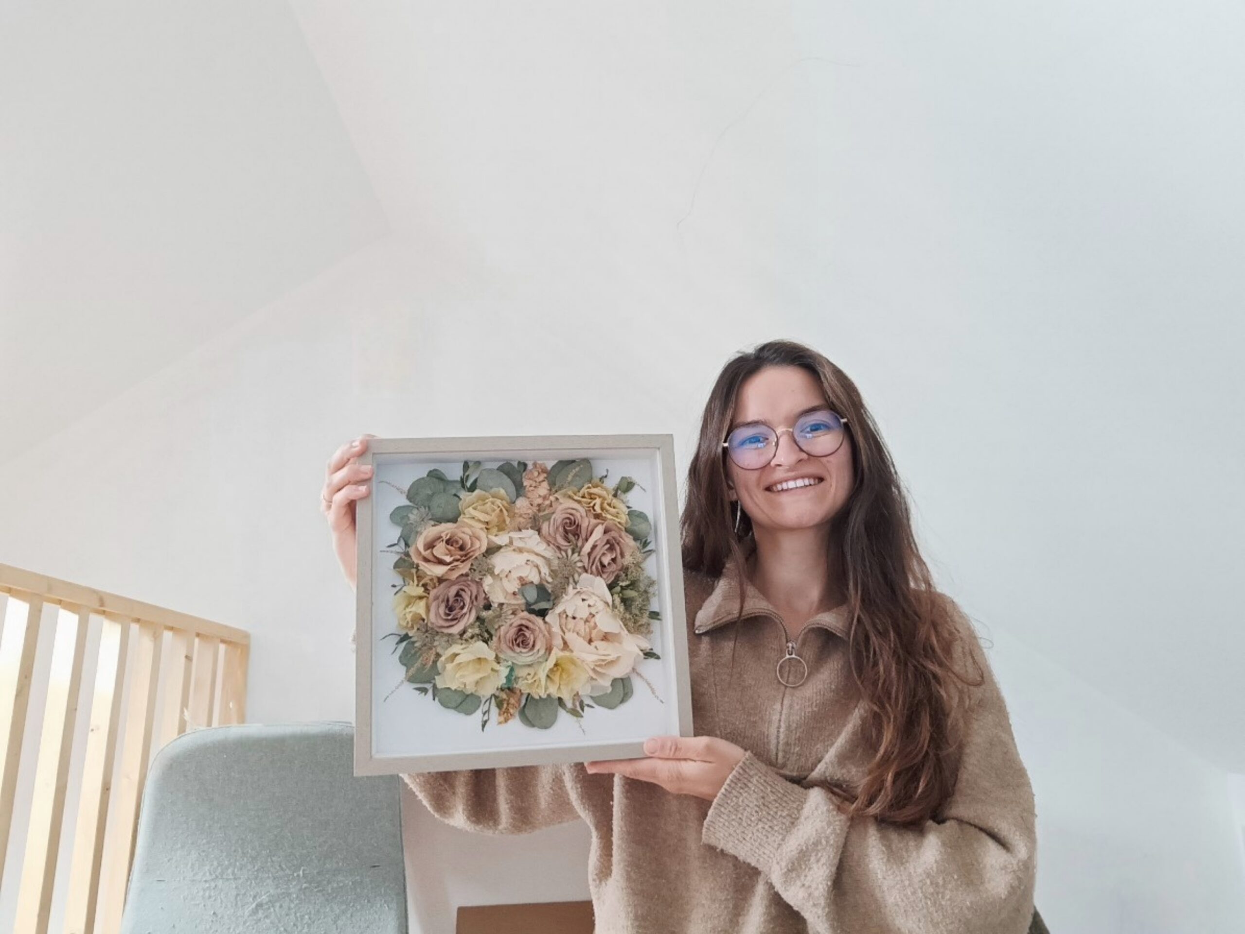 Artisane florale tenant un cadre contenant un bouquet de fleurs séchées préservées, souriante dans son atelier lumineux. Créatrice d'Invitation Florale basée en Normandie près de Caen